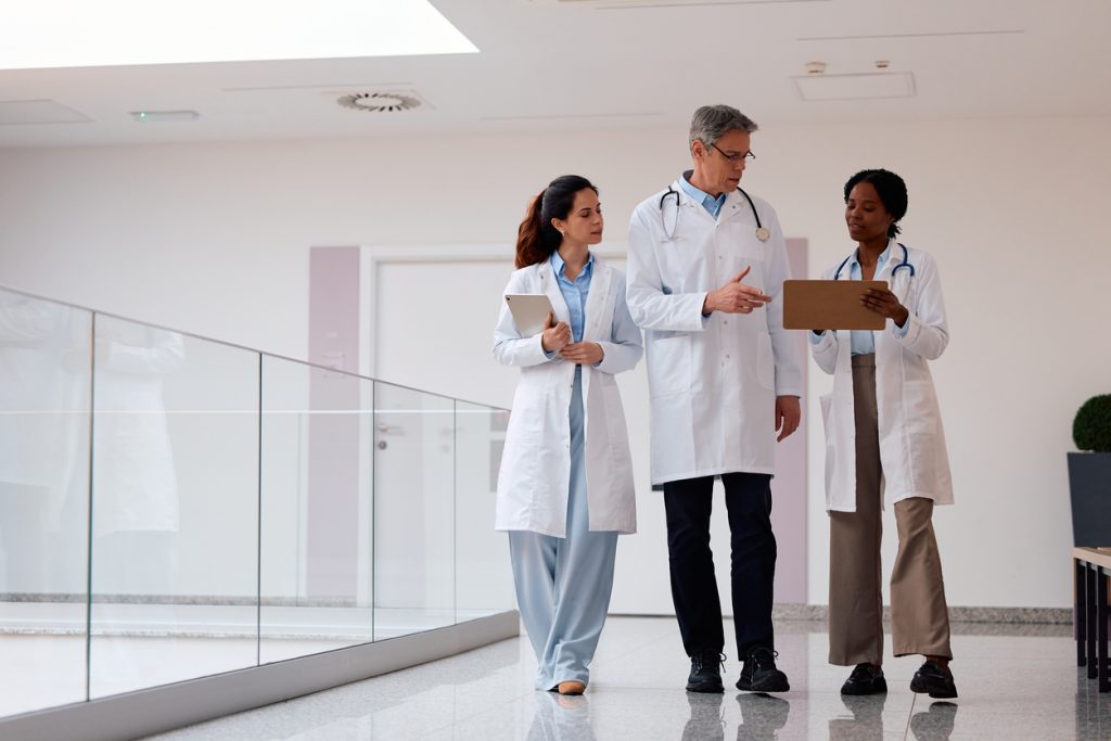 Diverse medical team of three doctors collaborating and discussing patient care while walking and reviewing documents in a modern hospital hallway. Emphasizing teamwork and professional communication