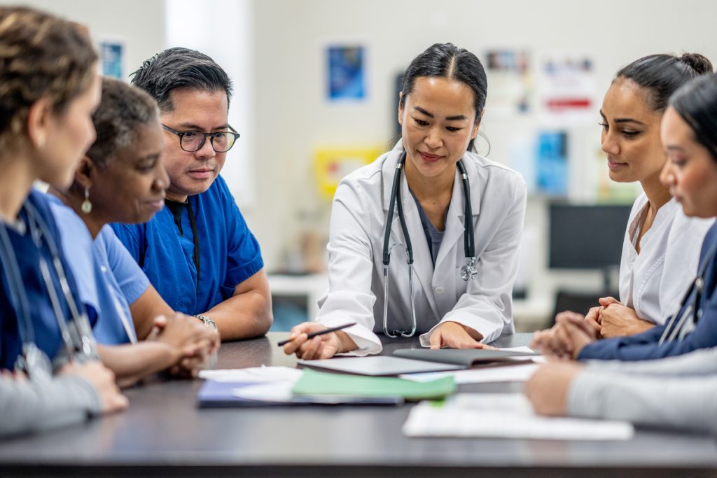 A small group of medical professionals, led by a doctor, are participating in team training as they sit around a table together.  They are each dressed professionally and are focused on the meetng.